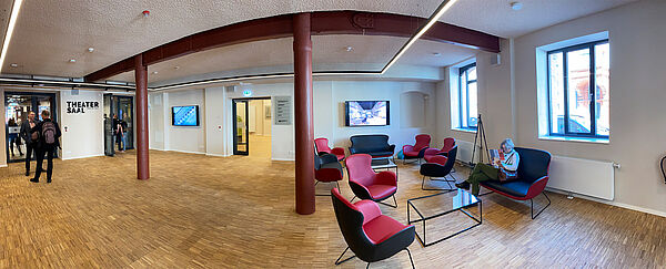Ein acht, neun Meter breiter Raum. Glastüren führen ins Restaurant und zum Treppenhaus des Theaters. An den Wänden hängen zwei Flachbildschirme. Der Boden trägt Holzoptik. Rechts im Raum stehen rot-schwarze Sessel.