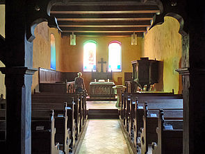 Links und rechts reihen sich Kirchenbänke. Am Ende des Raumes steht ein Altar. Dahinter befinden sich zwei bunt verglaste Fenster, durch die Sonnenlicht scheint.