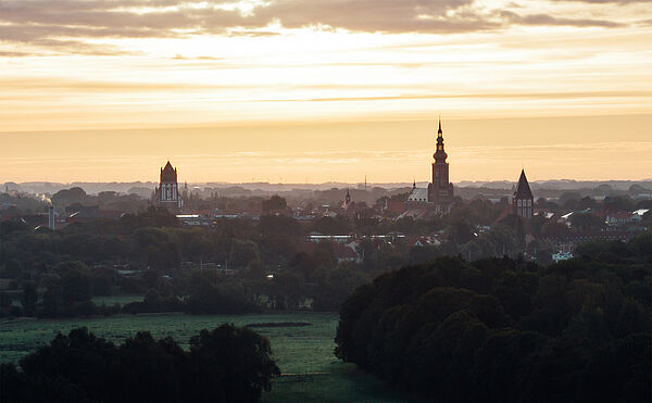 Ein orangefarbener Himmel bei Sonnenaufgang. Häuser, Bäume, Wiesen und Dächer bilden eine Silhouette, aus der Kirchentürme herausragen. 