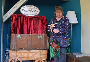 Martina Kriedel hält eine Hexen-Marionette in der Hand. Neben ihr steht auf einem Handwagen ein geöffneter Koffer. Sein Deckel ist mit rotem Samt ausgekleidet. Über ihm hängt ein Schild mit der Aufschrift "Koffertheater".