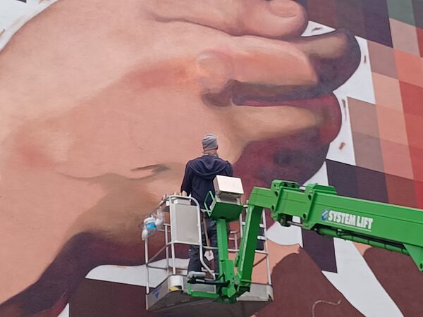 Ein Künstler steht auf einer Hebebühne und malt an einem Wandbild. Das Bild zeigt eine riesige Hand.