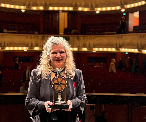 Eine Frau steht auf einer Bühne. Sie hält ihren Preis in der Hand. Im Hintergrund sind unscharf die beleuchteten Ränge vom Theater zu erkennen.