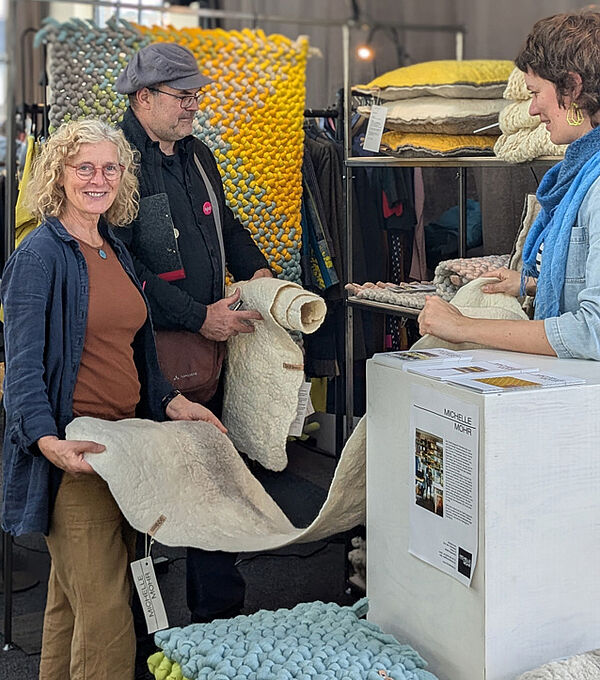 Ein Messestand. Eine Künstlerin zeigt zwei Besuchern ihre textile Kunst.