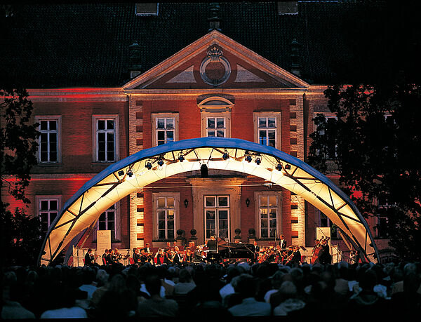 Vor der Backsteinfront von Schloss Bothmer steht eine Konzertbühne. Es ist dunkel, die Bühne leuchtet. Auf der Bühne spielen Musikerinnen und Musiker. Vor ihnen sitzt Publikum.