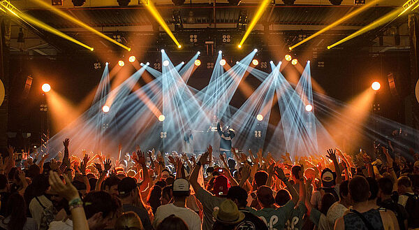 Eine Bühne. Auf der Bühne sind Musiker. Die Bühne ist bunt angeleuchtet. Vor der Bühne stehen viele Menschen. 