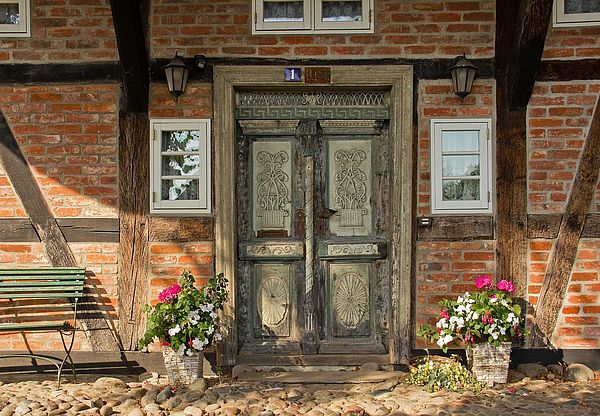 Ein Fachwerkhaus mit einer historischen Eingangstür. Davor stehen eine Bank und blühende Blumenkübel.