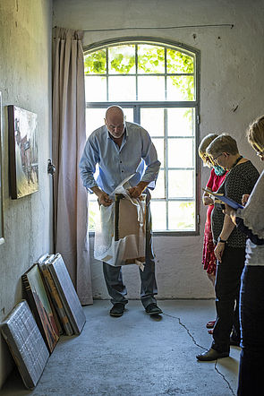 Jörg Herold nimmt einen Bilderrahmen aus einer Plastikfolie. An der Wand lehnen Leinwände. Davor stehen Mitglieder der Kunstkommission.