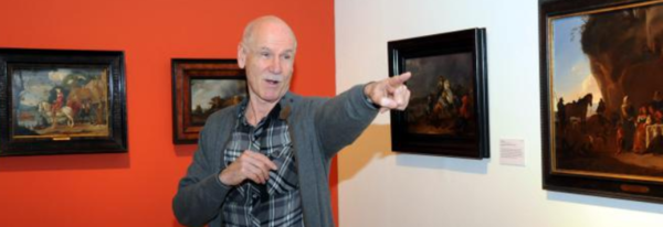 Christoph Müller steht vor einer Wand mit Gemälden und zeigt mit dem Finger in den Raum.