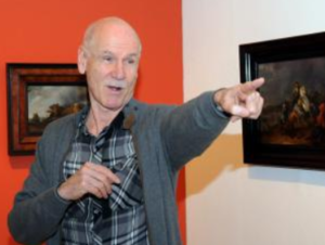 Christoph Müller steht vor einer Wand mit Gemälden und zeigt mit dem Finger in den Raum.
