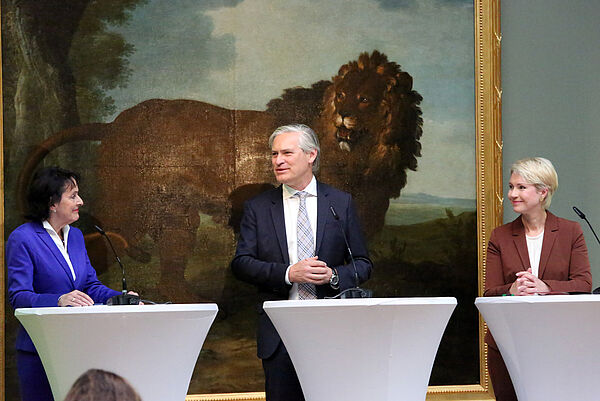 Drei Menschen an Stehpulten auf einer Bühne.