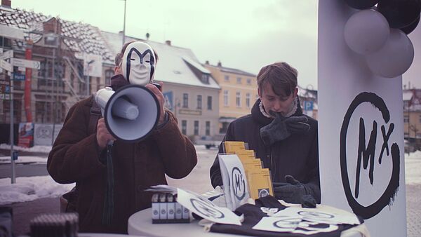 Eine Filmszene zeigt zwei junge Männer auf einerStraße. Der Linke trägt eine Maske und ein Megaphone. Der Recht wühlt in Papierzetteln.