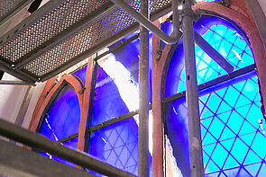 Blick auf ein Gerüst. Dahinter schimmert ein Fenster blau.