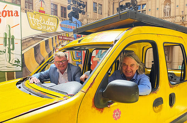 Geschäftsführer Christian Schwandt und Generalintendant Hans-Georg Wegner sitzen auf der Bühne im Schweriner Schlossinnenhof in einem gelben Auto. 
