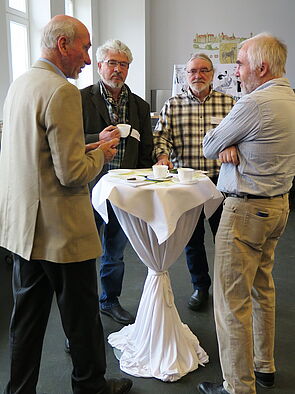 Vier Männer stehen in der Pause an einem Bistro-Tisch und unterhalten sich.