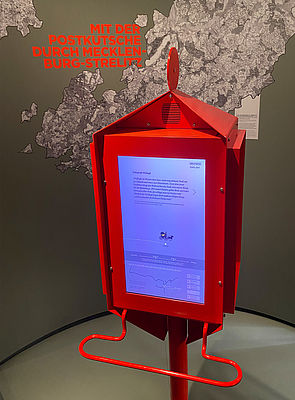 In einen roten Postkasten ist ein Bildschirm integriert. Er zeigt an, auf welchen Wegen man früher von Neustrelitz mit der Postkutsche reisen konnte.