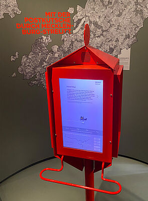 In einen roten Postkasten ist ein Bildschirm integriert. Er zeigt an, auf welchen Wegen man früher von Neustrelitz mit der Postkutsche reisen konnte.