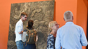Vier Besucher stehen vor einem Bild, das etwa zwei Meter hoch an der Wand lehnt.