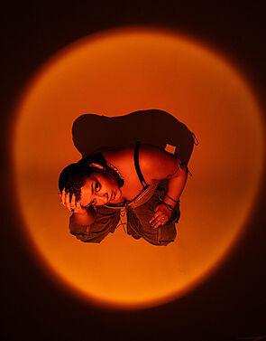 Eine Frau hockt in einem orangefarbenen Lichtkreis. Sie schaut nach oben. 