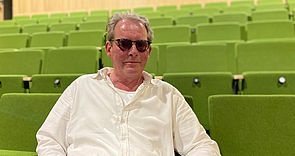 Thomas Ott-Albrecht sitzt auf einem der grünen Theatersessel. Thomas Ott-Albrecht ist Anfang 60, trägt ein weißes Hemd und eine Sonnenbrille. 