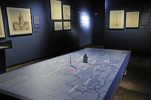 Im Raum steht ein Tisch mit einer Karte von Europa. An den blauen Wänden hängen angeleuchtete Gemälde.