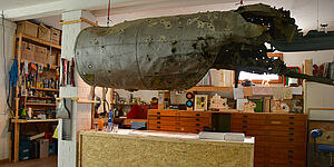Ein Hubschraubertank hängt von der Decke. Im Atelier stehen Regale und ein Schreibtisch mit vielen Schubladen. Überall stehen und liegen Arbeitsutensilien. 