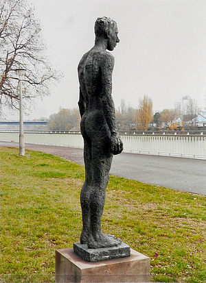 Skulptur eines nackten Mannes von der Seite. Er steht aufrecht mit geschlossenen Beinen. Die Arme hängen links und rechts herab.