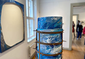 Eine blau bemalte Rolle steht zusammengerollt im Raum und wird von einem Eisengitter umgeben. An der Wand hängt eine Leinwand mit einem Loch.