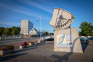Ein Straße führt auf eine Brücke. Eine Skulptur steht vor der Brücke.
