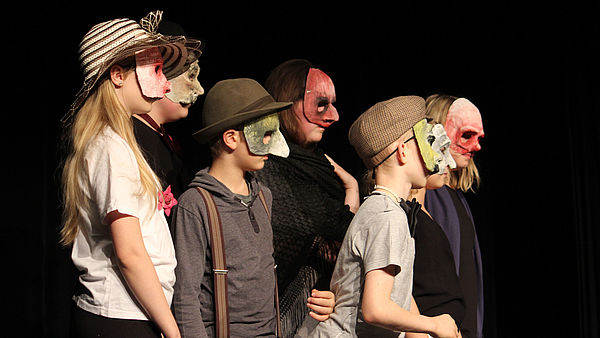 Sieben Kinder stehen mit Masken, die über Augen und Nase reichen, auf einer Bühne.