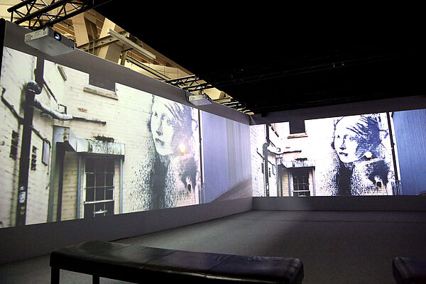 Ein Ausstellungsraum. Das Bild zeigt eine Hauswand. Auf der Hauswand ist ein Bild von einem Mädchen.