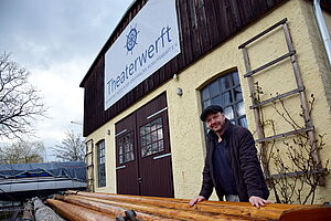 Jens Hasselmann steht draußen, hinter der Theaterwerft. Vor ihm liegen lange Holzbalken. Hinter ihm steht die Theaterwerft, ein gelbes Gebäude mit dunklem Holz.