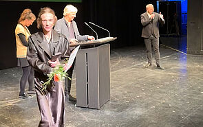 Eine Frau geht von einer Bühne. Sie hat Blumen und einen Preis in der Hand. Im Hintergrund stehen Menschen auf der Bühne. 