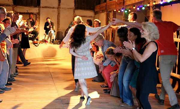 Eine Scheune. Frauen und Männer tanzen und klatschen. Im Hintergrund spielen Musiker.