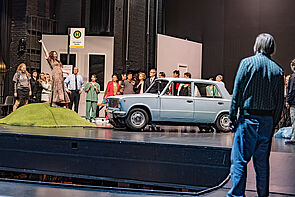 Schauspieler auf der Bühne. Vor ihnen steht ein blaues Auto.