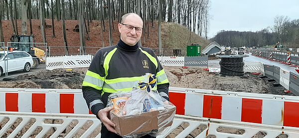 Wernfried Heinz Müller steht mit einem Präsentkorb in der Hand vor einer abgesperrten Baustelle. 