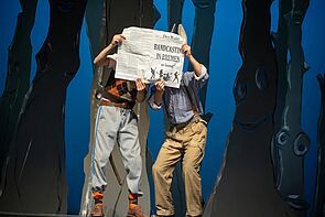 Zwei verkleidete Menschen stehen auf einer Theaterbühne und halten sich eine Zeitung vor das Gesicht.