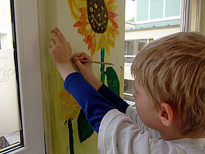 Ein Kind malt mit einem Stift an eine Wand, auf der sich bereits eine Sonnenblume und andere Blumen befinden.