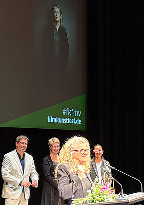 Eine Frau steht an einem Rednerpult und spricht. Auf dem Rednerpult steht ein Preis. Daneben liegen Blumen. Hinter der Frau stehen drei Leute: der Festivalleiter, die Ministerpräsidentin und die Laudatorin.
