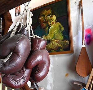 An einem Haken hängen alte Boxhandschuhe. Dahinter hängt ein Gemälde. Das Gemälde zeigt einen Mann mit einem Kind.