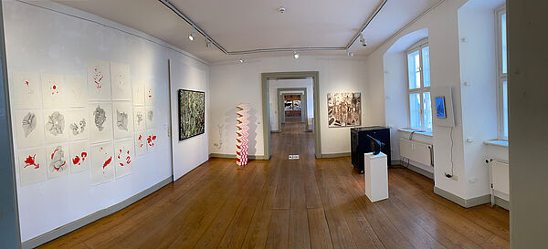 Ein Ausstellungsraum. An der Wand hängen Bilder. Im Raum steht eine Plastik.