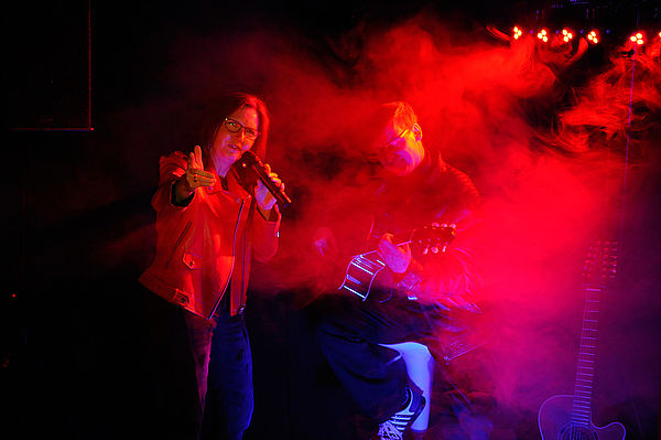 Eine Bühne. Im roten Scheinwerferlicht liegt Nebel. Auf der Bühne stehen Andrea Peters und Thomas Birkigt. Sie hat ein Mikrofon in der Hand, er eine Gitarre.