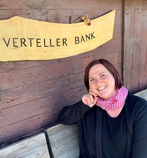 Eine Frau sitzt auf einer Bank. Die Frau ist Johanna Bojarra. Hinter der Bank hängt ein Schild. Auf dem Schild steht das plattdeutsche Wort Verteller Bank.