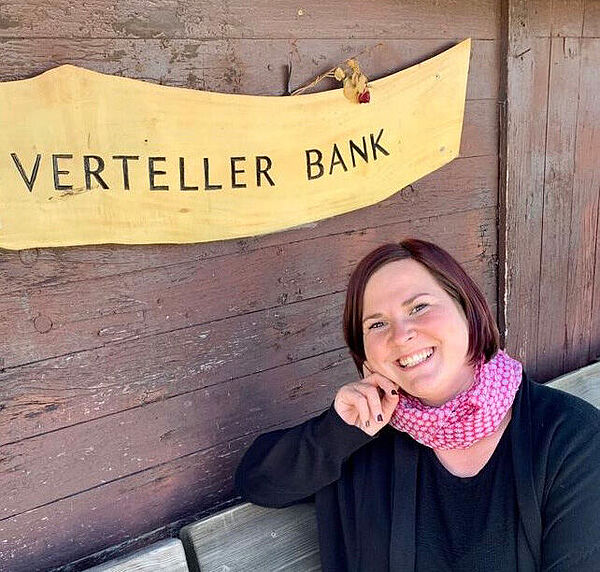 Eine Frau sitzt auf einer Bank. Die Frau ist Johanna Bojarra. Hinter der Bank hängt ein Schild. Auf dem Schild steht das plattdeutsche Wort Verteller Bank.