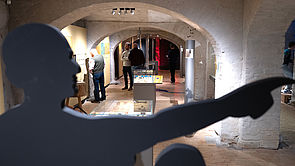 Im Vordergrund ein Schattenschnitt von einer Figur, die nach rechts zeigt. Im Hintergrund eine Ausstellungshalle.