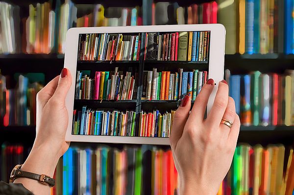 Eine Frau hält ein Tablet vor ein gefülltes Bücherregal, so dass ein Ausschnitt des Regals wie ein Foto im Tablet abgebildet wird.
