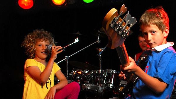 Ein Mädchen steht an einem Mikrofon. Ein Junge hat eine Gitarre in der Hand.