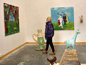 Eine Frau im Ausstellungsraum betrachtet ein Gemälde. Im Vordergrund mehrere Skulpturen.