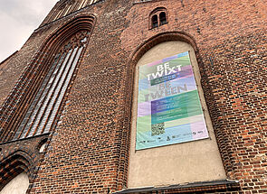 An der Außenfassade vom Kircheneingang hängt ein großes Plakat, das auf die Landesweite Kunstschau hinweist. 