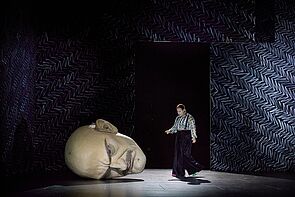 Ein großer Kopf liegt auf der Bühne. Daneben singt ein Schauspieler.