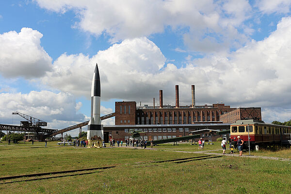 Vor dem Kraftwerk, einem Backsteinbau, steht ein Zug und eine Rakete. Besucherinnen und Besucher spazieren über das Außengelände.